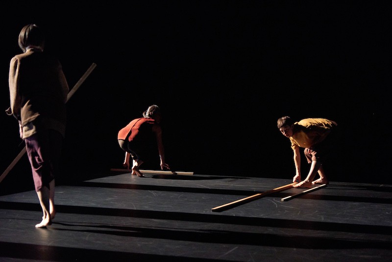 Three women in semi darkness hold sticks or lay down the long wooden rods. They cast long shadows.